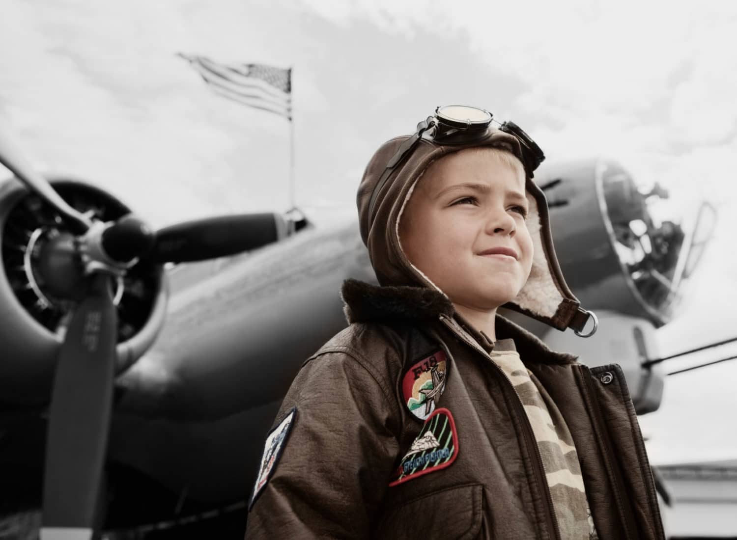 young boy in flying hat and goggles standing in front of aeroplane